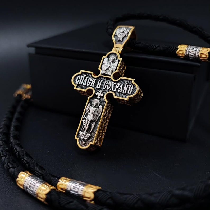 May include: A silver and gold-toned pendant in the shape of a cross, with intricate religious iconography and the inscription "Спаси и Сохрани". The pendant hangs on a black braided cord with gold and silver accents.