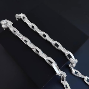 May include: Two silver-colored chain necklaces with a geometric link design. The chains are displayed on a black surface, showcasing their reflective quality. The links have a unique, angular shape, creating a modern aesthetic.