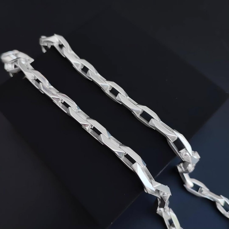 May include: Two silver-colored chain necklaces with a geometric link design. The chains are displayed on a black surface, showcasing their reflective quality. The links have a unique, angular shape, creating a modern aesthetic.