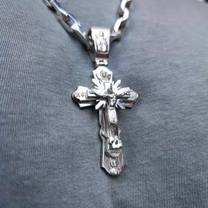 May include: A silver-colored Orthodox cross pendant with a detailed depiction of Jesus Christ on the cross. The pendant is attached to a thick, silver chain. The cross has intricate carvings and is set against a gray background.