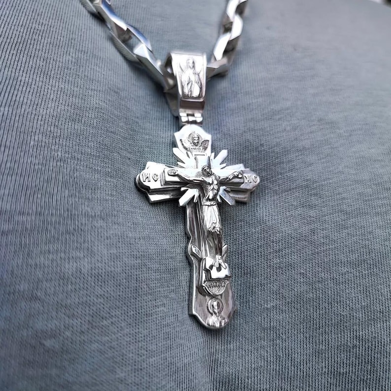 May include: A silver-colored Orthodox cross pendant with a detailed depiction of Jesus Christ on the cross. The pendant is attached to a thick, silver chain. The cross has intricate carvings and is set against a gray background.