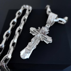 May include: A silver-colored Orthodox cross pendant with intricate detailing, featuring a depiction of Jesus Christ. The cross is displayed next to a matching silver chain, both reflecting light. The design is ornate and detailed.
