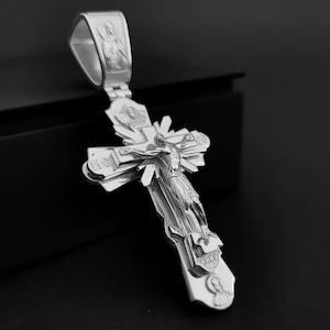 May include: A silver pendant in the shape of a cross, featuring a detailed depiction of Jesus Christ on the cross. The pendant has intricate engravings and religious iconography, with a loop at the top for a chain. The cross is set against a dark background.