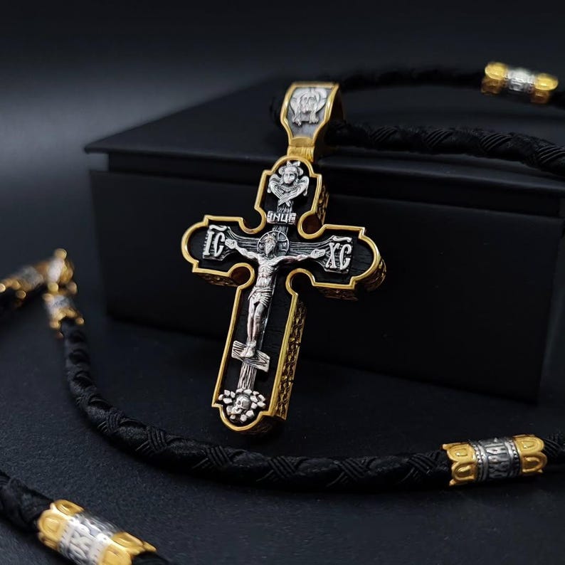 May include: A silver and gold-toned Orthodox cross pendant with a black background. The cross features a detailed depiction of the crucifixion, with ornate detailing and a braided black cord. The pendant has gold accents and is likely made of metal.