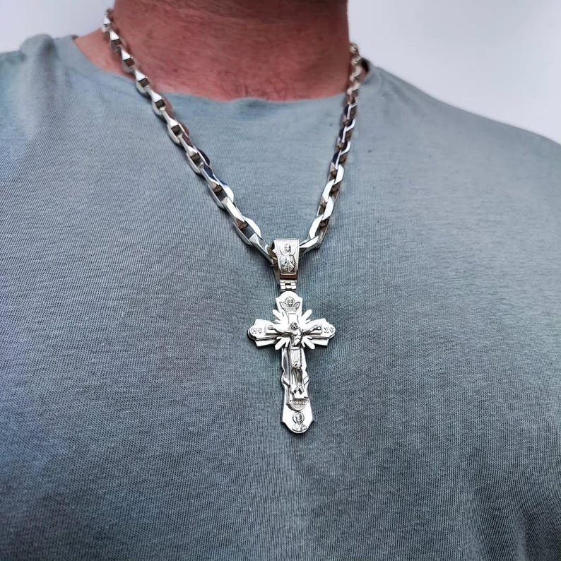 May include: A silver-colored pendant necklace featuring a detailed cross design. The cross has intricate carvings and is suspended from a thick, linked chain. The necklace is displayed against a gray background.