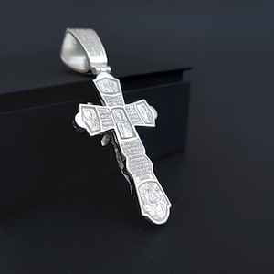 May include: A silver pendant in the shape of a cross. The cross features intricate engravings of religious figures and text. The pendant has a loop at the top for a chain and is displayed against a dark background.