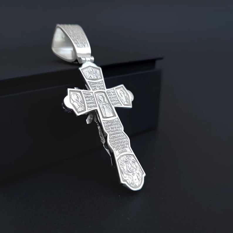 May include: A silver pendant in the shape of a cross. The cross features intricate engravings of religious figures and text. The pendant has a loop at the top for a chain and is displayed against a dark background.