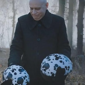 Puede incluir: Dos cascos blancos con manchas negras son sostenidos por una persona que lleva un abrigo negro. Los cascos tienen forma redondeada y un acabado brillante. El fondo es una escena de bosque borrosa.