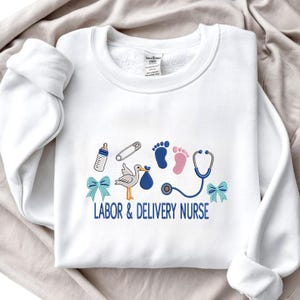 May include: White sweatshirt with embroidered baby-themed designs. Features a baby bottle, safety pin, footprints, stork carrying a bundle, stethoscope, and blue bows. The text "LABOR & DELIVERY NURSE" is embroidered in blue.