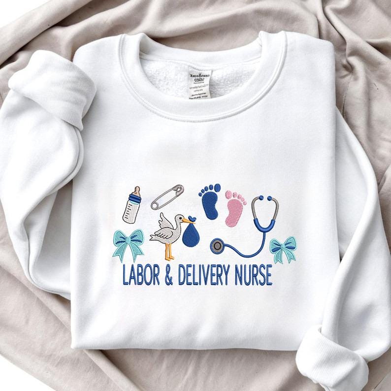 May include: White sweatshirt with embroidered baby-themed designs. Features a baby bottle, safety pin, footprints, stork carrying a bundle, stethoscope, and blue bows. The text "LABOR & DELIVERY NURSE" is embroidered in blue.