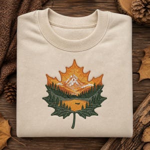 Op de afbeelding: Crèmekleurige sweatshirt met een gedetailleerd geborduurd ontwerp van een berglandschap in een esdoornblad. Het ontwerp toont een bergketen, bomen en een meer, met oranje, groene en witte kleuren. De sweatshirt heeft een ronde hals en geribde manchetten.