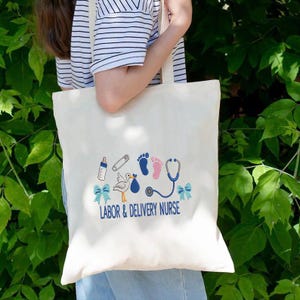 May include: Cream-colored tote bag with a blue and pink baby-themed design. The design includes a baby bottle, footprints, a stork, and a stethoscope. The text "LABOR & DELIVERY NURSE" is printed in blue.