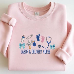 May include: A light pink sweatshirt with embroidered baby-themed graphics. The design includes a baby bottle, safety pin, footprints, stork, stethoscope, and bows. The text "LABOR & DELIVERY NURSE" is embroidered in blue.
