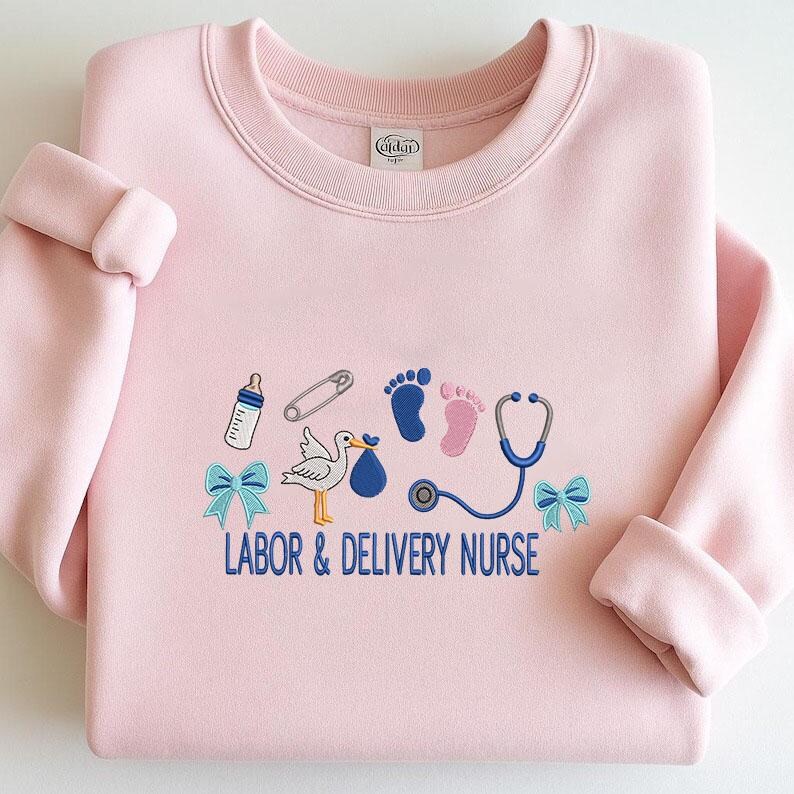 May include: A light pink sweatshirt with embroidered baby-themed graphics. The design includes a baby bottle, safety pin, footprints, stork, stethoscope, and bows. The text "LABOR & DELIVERY NURSE" is embroidered in blue.