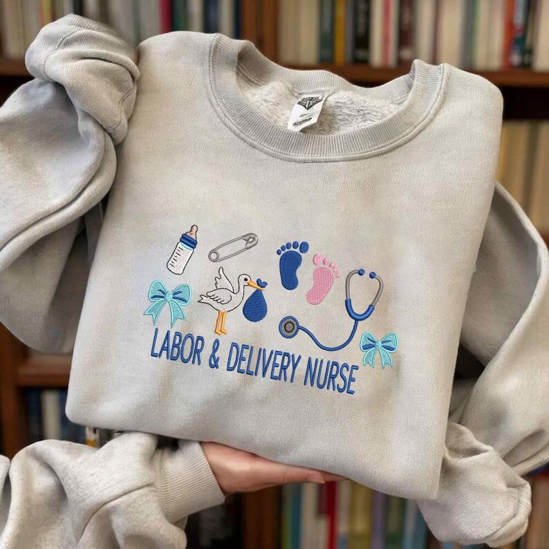May include: A light gray sweatshirt with embroidered baby-themed designs, including a stork, baby feet, a bottle, and a stethoscope. The text "LABOR & DELIVERY NURSE" is embroidered in blue. The sweatshirt has a crew neck and long sleeves.