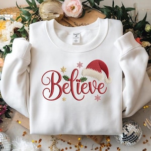 Peut inclure: Un sweat-shirt blanc avec le mot "Believe" brodé en rouge, orné d'un bonnet de Père Noël et d'éléments festifs. Le motif comprend des étoiles dorées, des flocons de neige et du houx, créant une ambiance de Noël.