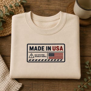 Può includere: Felpa color crema con girocollo. Una toppa rettangolare mostra il testo "MADE IN USA" in blu navy e rosso, insieme a una bandiera americana e le frasi "BUILT WITH PRIDE" e "DESIGNED IN AMERICA".