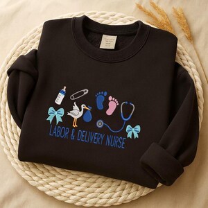 May include: Black sweatshirt with embroidered baby-themed designs, including a stork, footprints, and a stethoscope. The text "LABOR & DELIVERY NURSE" is embroidered in blue. The sweatshirt has a crew neck and long sleeves.