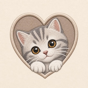 Puede incluir: Un parche bordado que presenta un gatito atigrado gris y blanco con grandes ojos color ámbar, posicionado dentro de un marco en forma de corazón con fondo marrón claro. El gatito tiene bigotes visibles y sus patas delanteras descansan.