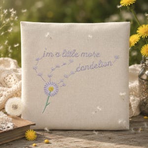 May include: A square, off-white pillow with embroidered text and a dandelion design. The text reads "I'm a little more dandelion" in light purple thread. The dandelion has a purple and white flower head and a green stem. The pillow is on a wooden surface.