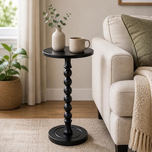 May include: A black side table with a round top and base, connected by a series of spherical accents. A beige mug and a small vase with greenery sit on top. The table is next to a beige sofa with a green pillow.
