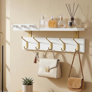 May include: A white wall-mounted shelf with a row of hooks and a top shelf. The hooks are gold-colored, and the shelf holds a purse, keys, and decorative items. A small succulent plant in a white pot sits on a wooden bench.