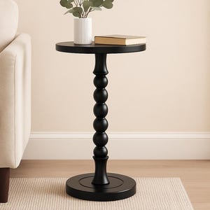 May include: A black side table with a round top and a decorative, beaded column. A small white vase with greenery and a closed book rest on the table. The table is approximately 24 inches tall.