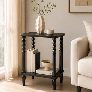May include: A black two-tier side table with four spindle-style legs. The top surface holds a white wavy vase with green stems and a small ribbed bowl. The bottom shelf contains three upright books and a shallow beige bowl resting on a book.