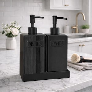 Op de afbeelding: Zwarte keramische zeepdispenser set voor keuken of badkamer. Eén dispenser is gemarkeerd met "DISHES" en de andere met "HANDS". Beide hebben zwarte pompknoppen. De dispensers staan op een rechthoekige basis.
