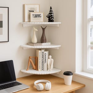 May include: A corner shelf unit with three white shelves, each adorned with a beaded edge. The shelves display decorative items like vases, books, and framed art. A laptop, headphones, and a coffee cup sit on the desk below.