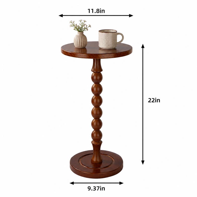Op de afbeelding: Een ronde bruine houten bijzettafel met een spilvormige voet. De tafel heeft een diameter van 29,97 cm aan de bovenkant, 23,8 cm aan de basis en een hoogte van 55,88 cm. Een kleine witte vaas met bloemen en een mok staan op het oppervlak.