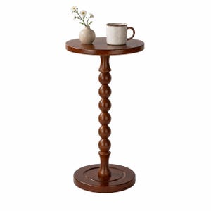 Op de afbeelding: Een ronde bijzettafel van donkerbruin hout met een centrale kolom bestaande uit gestapelde bollen. Op het tafelblad staat een kleine beige keramieken vaas met drie witte bloemen en een gespikkelde beige keramieken mok met een bruine rand.