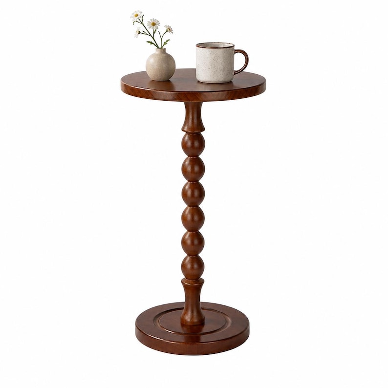 Op de afbeelding: Een ronde bijzettafel van donkerbruin hout met een centrale kolom bestaande uit gestapelde bollen. Op het tafelblad staat een kleine beige keramieken vaas met drie witte bloemen en een gespikkelde beige keramieken mok met een bruine rand.