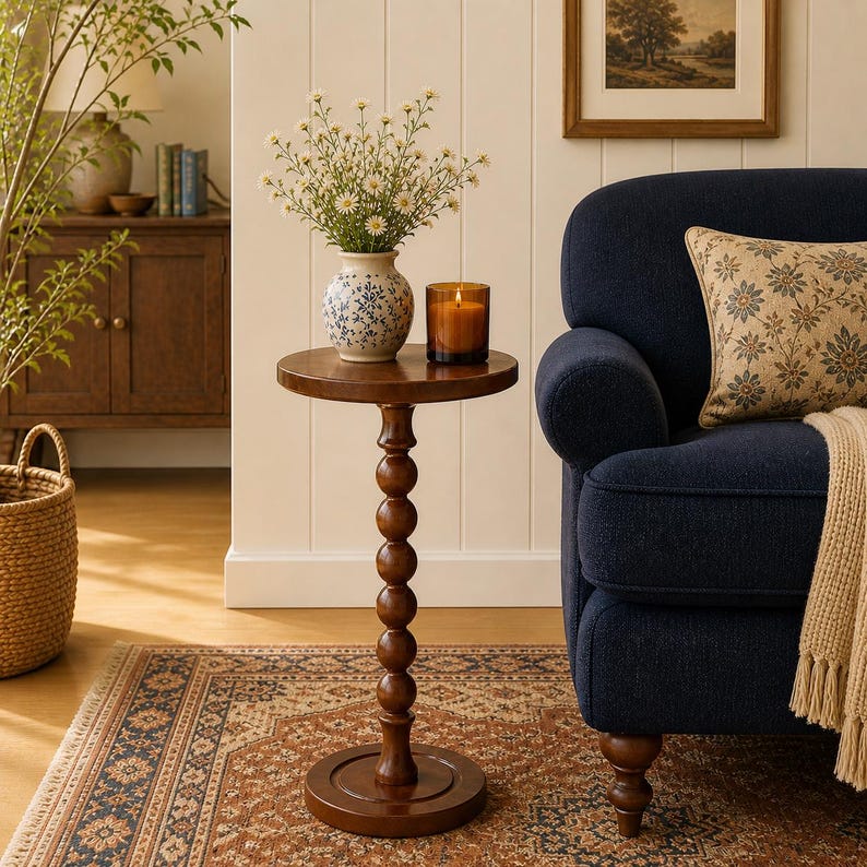 Op de afbeelding: Een ronde houten bijzettafel met een centrale kolom bestaande uit gestapelde bollen. De tafel heeft een donkerbruine afwerking en een ronde voet. Er staat een witte keramische vaas met blauwe bloemmotieven en kleine witte bloemen op, plus een kaars.