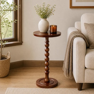 May include: A dark brown, round side table with a turned, beaded column base. A white vase with flowers and a lit candle sit on top. The table is next to a cream-colored sofa with a brown throw blanket.