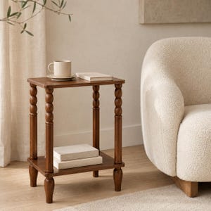 May include: A square wooden side table with four turned, spindle-style legs and a lower shelf. The table features a smooth, dark brown finish. A plain white ceramic mug sits on the top surface next to a closed, light-colored hardcover book.