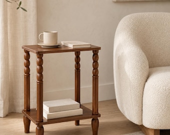 May include: A square wooden side table with four turned, spindle-style legs and a lower shelf. The table features a smooth, dark brown finish. A plain white ceramic mug sits on the top surface next to a closed, light-colored hardcover book.