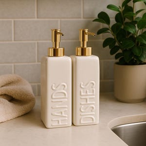 May include: Two off-white ceramic soap dispensers with gold-colored pump tops. One dispenser reads "HANDS" and the other reads "DISHES" in embossed lettering. The dispensers are rectangular and sit on a white countertop.