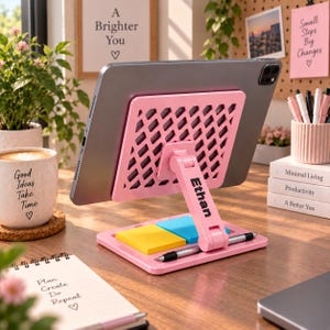 May include: A pink adjustable tablet stand with a lattice-patterned back panel and the name Ethan printed on the support arm. The base features a recessed area holding yellow and blue sticky notes and a black pen resting horizontally in a groove.