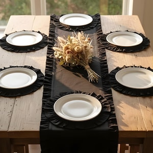 May include: A dining table set with a black table runner and matching placemats. White plates are arranged on the placemats. A bouquet of dried flowers sits in the center of the table. The table is made of wood.