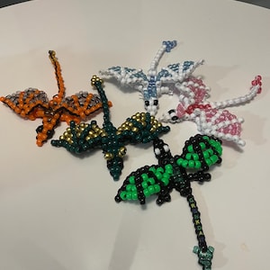 May include: A selection of beaded dragon figurines in various colours, including orange, green, blue, and pink. The dragons have detailed wings and bodies, with some featuring gold accents. The figurines are displayed on a white surface.