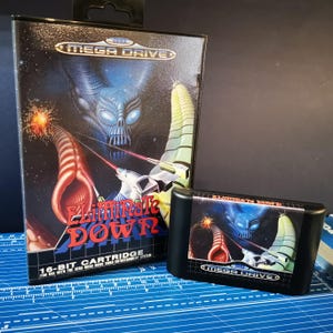 May include: A black Mega Drive video game cartridge and box set for the game "Eliminate Down." The box art shows a space battle scene with a blue alien and red laser beams. The cartridge is black with the game title.