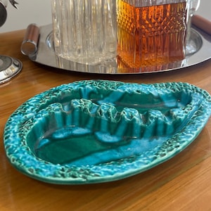 May include: An oval-shaped ceramic ashtray featuring a vibrant turquoise and teal glaze with a textured, craggy rim. The interior basin includes three molded notches for resting items, finished with a glossy, multi-tonal green surface.
