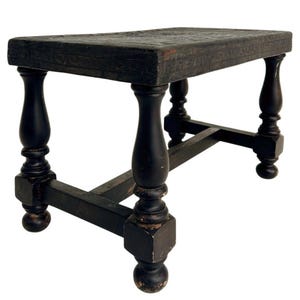 May include: A black wooden bench with a rectangular top featuring a carved Greek key pattern along the edges. The bench stands on four turned legs connected by a lower H-shaped stretcher, showing signs of wear and distressed paint finish.