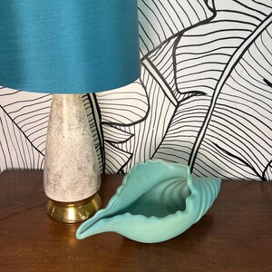 May include: A teal seashell-shaped ceramic dish sits on a wooden surface next to a lamp with a teal lampshade and a speckled cream-colored base with a gold-colored base. The background features a black and white leaf pattern.