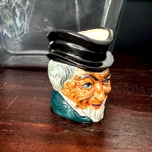 May include: A ceramic character jug shaped like an elderly person wearing a black top hat. The figure has a tan face, grey hair, a white goatee, and a dark green coat collar. The interior of the vessel is cream-colored, and the exterior features detailed facial expressions with painted blue eyes and red lips.