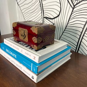 May include: A small, rectangular, red wooden box with brass hardware and a decorative clasp sits atop a stack of three books. The books have white and blue covers with titles like "BEAUTY BY DESIGN" and "DECORATE". The background features a black and white leaf pattern.