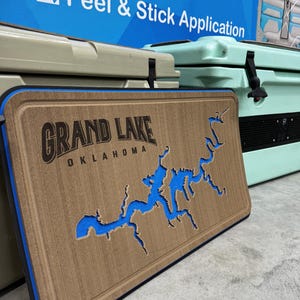Peut inclure: Planche à découper en bois avec une carte détaillée du lac Grand, Oklahoma, avec des détails bleus représentant l'eau. Le texte "GRAND LAKE OKLAHOMA" est visible. La planche a une surface marron avec un bord bleu.