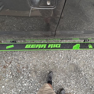May include: Black and green "BEAR RIG" running board attached to a gray vehicle. The running board has a diamond plate pattern with green text, a bear graphic, and bear paw prints.