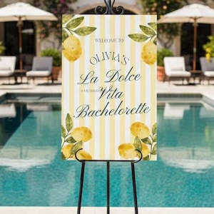 Może przedstawiać: Znak powitalny na wieczór panieński La Dolce Vita Olivii. Znak ma żółto-białe paski w tle z akwarelowymi cytrynami i liśćmi. Znak jest wyświetlany na czarnym metalowym sztaludze w pobliżu basenu.